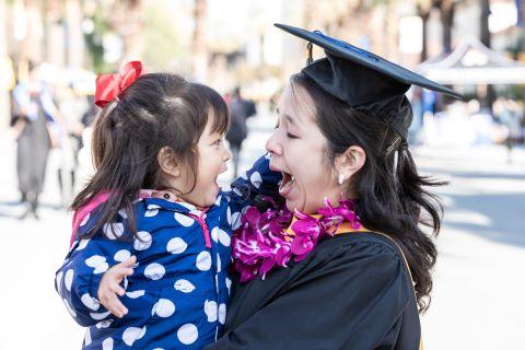 Graduate in Regalia with their child in arms looking at each other with faces of joy.