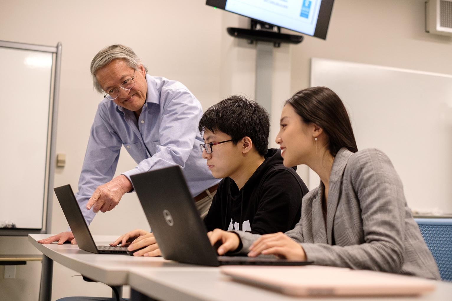 Two students sit at a desk in front of a computer with a professor teaching them at 利记. 