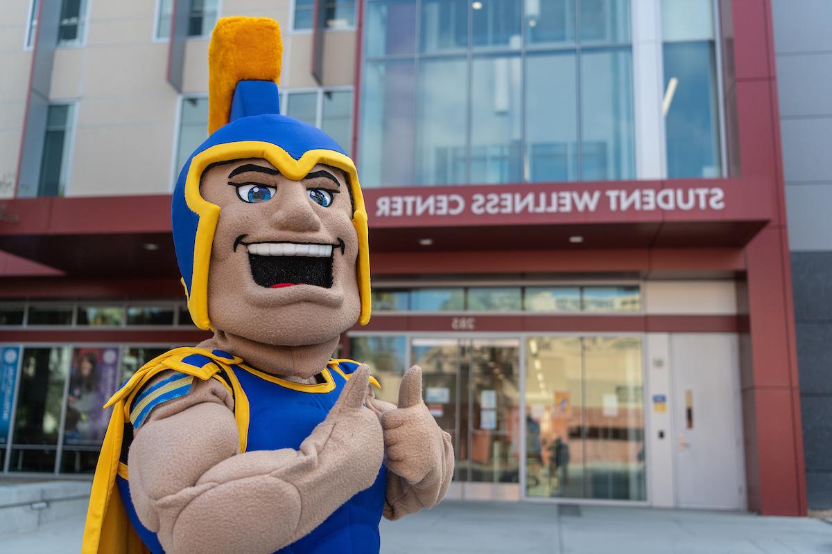 Sammy with their thumbs up standing in front of the Student Wellness Center
